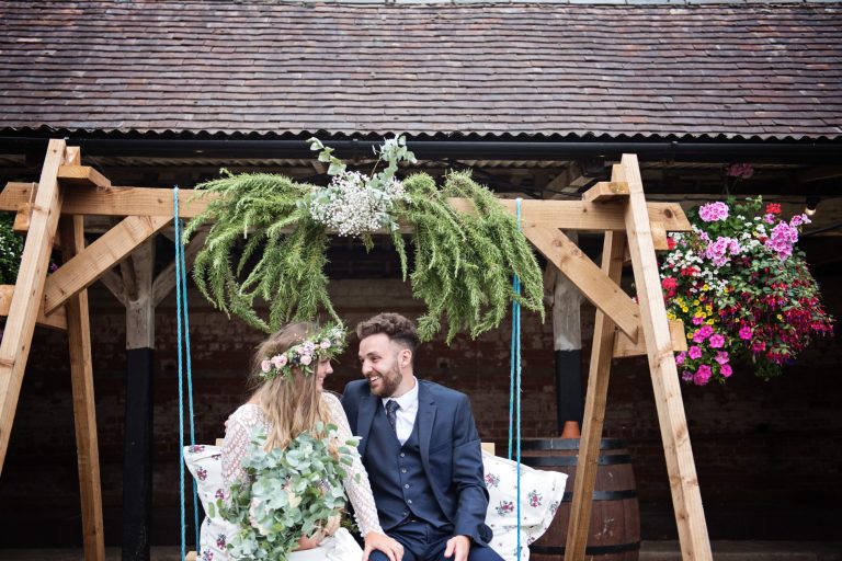 Bride and groom sitting and smiling as they sit on a wooden swing. Photo by Blooming Photography