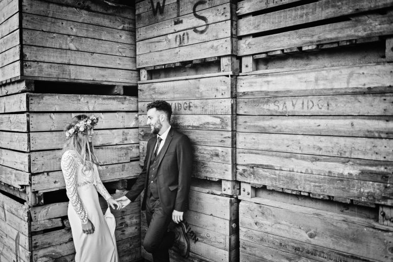 Rustic photo of bride and groom holding hands amongst a load of wooden box's. Photo by Blooming Photography