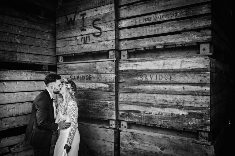 Rustic B&W photo of bride and groom kissing amongst a load of wooden box's. Photo by Blooming Photography