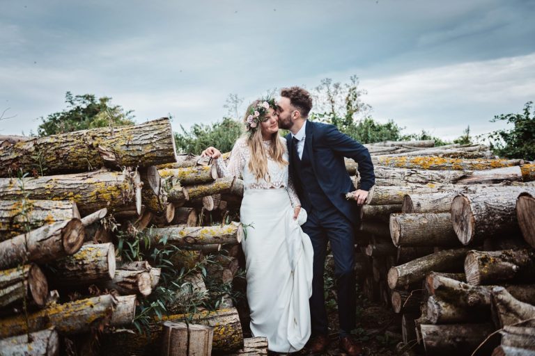 Dramatic photo of bride and groom kissing amongst a stack of logs. Photo by Blooming Photography