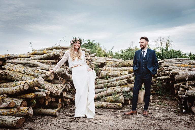 Dramatic photo of bride and groom standing amongst a stack of logs. Photo by Blooming Photography