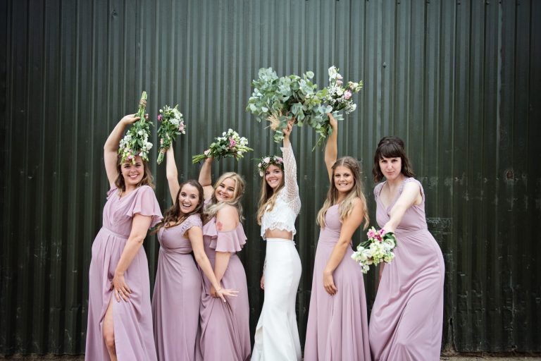 Bride and bridesmaids pose with their wedding flower bouquets held up in the air as they pose in front of an agricultural building.