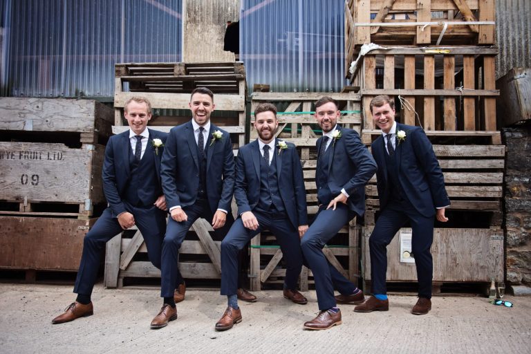 Groomsmen pull a lunge for the wedding photograph as they stand in front of wooden fruit pallets.