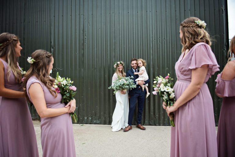 Bride and groom and their daughter stand together as their bridesmaids look at them. Candid fun photo by Gloucestershire Wedding Photographer, Blooming Photography