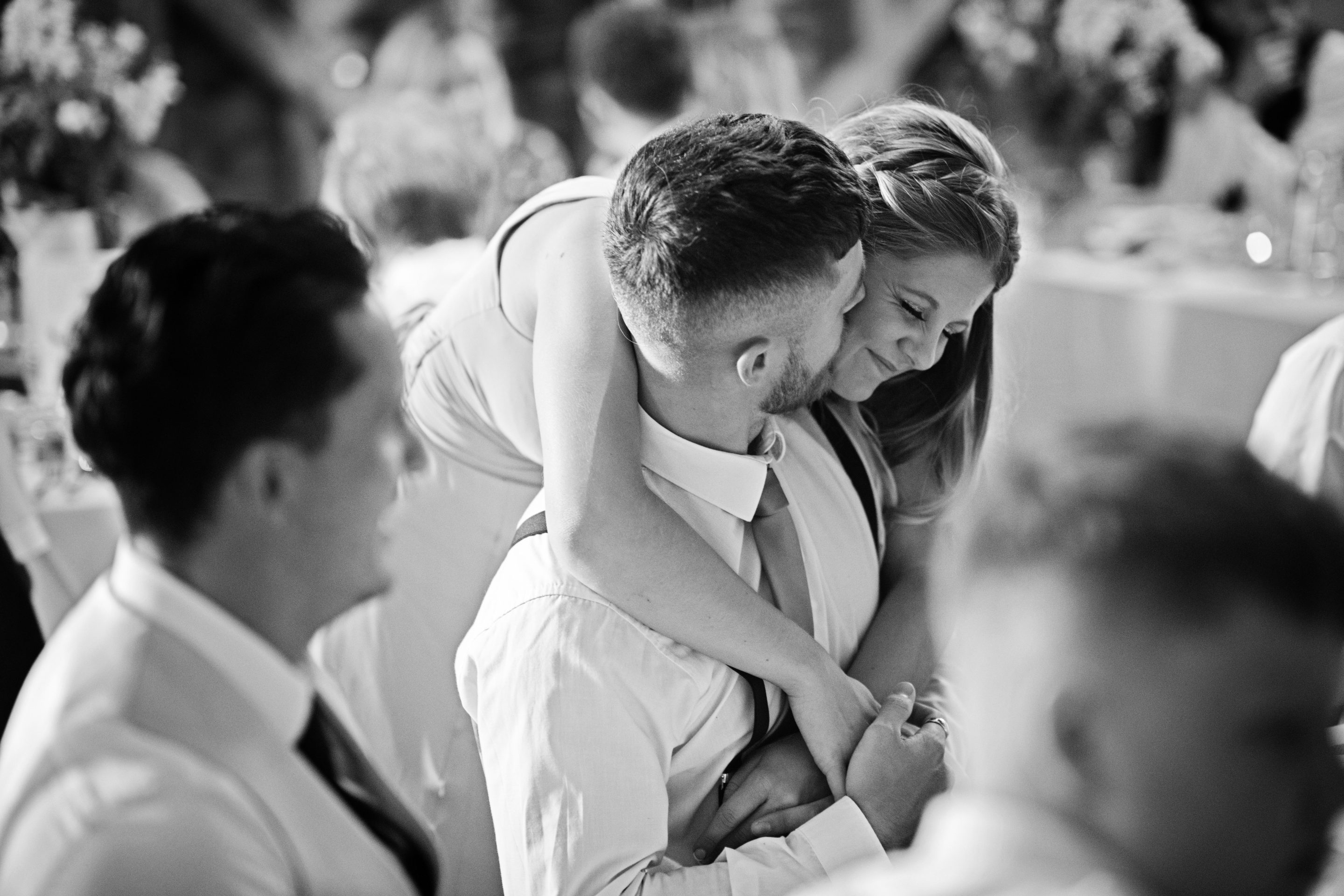Candid b&w photo of a bridemaids hugging a partner who is sitting down. At Overbarn wedding venue in Gloucester.
