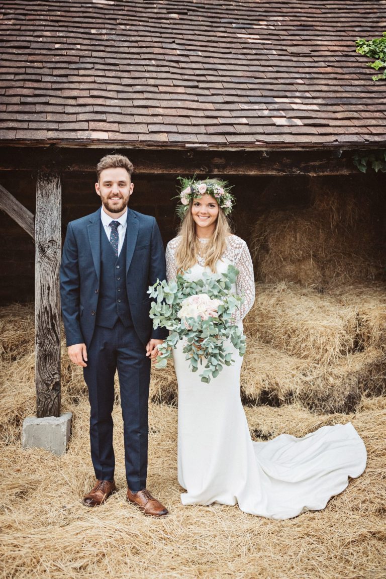 Bride and groom stand face on as they stand on a bed of straw. Photo by Blooming Photography