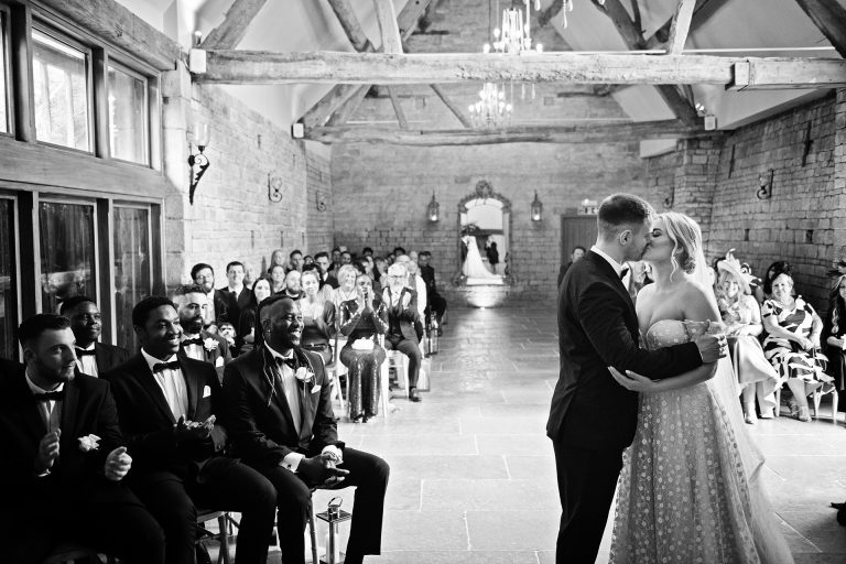 B&W image of bride and groom kissing as they have been announced husband and wife. Their guests celebrate behind them. Candid photo by Blooming Photography taken at Blackwell Grange.