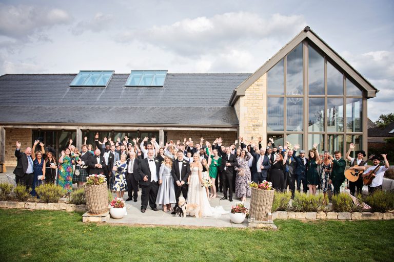 A large group photo of everyone at a wedding smiling as they put their hands in the air like they don't care! Photo by Blooming Photography taken at Blackwell Grange, Warwickshire wedding.