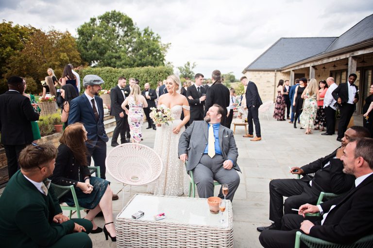 Candid photo by Blooming Photography taken at Blackwell Grange, Warwickshire wedding. A bride chats to her guests, some of them are smoking but they are all smiling