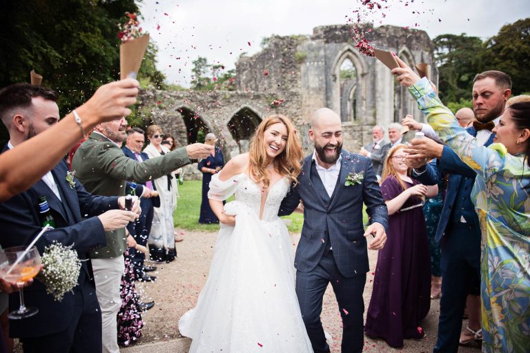 Colour photo of a bride and groom smiling whilst having wedding confetti thrown at them at Margam Country Park..