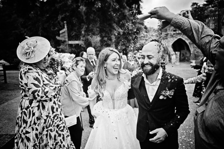 Natural black & white photo of a bride and groom smiling whilst having wedding confetti thrown at them.