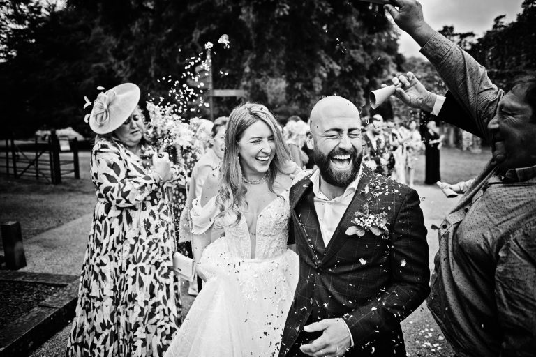 Black & white photo of a bride and groom smiling whilst having wedding confetti thrown at them at Margam