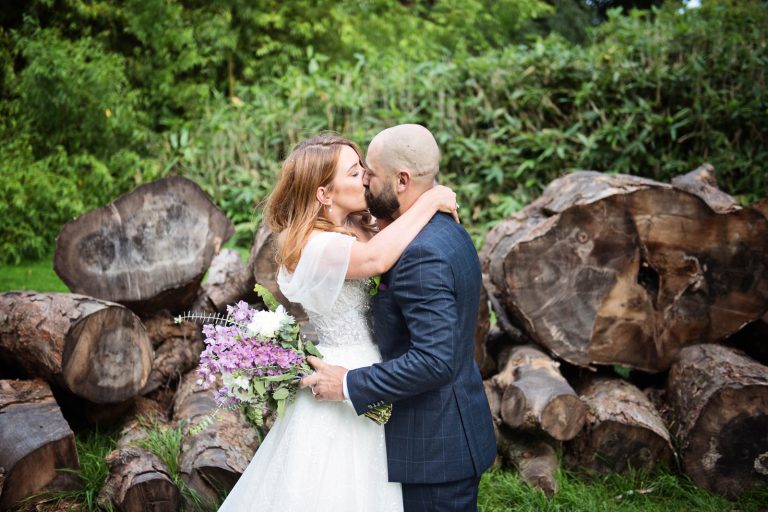 Photo of bride and groom holding each other closely, kissing with logs behind them.