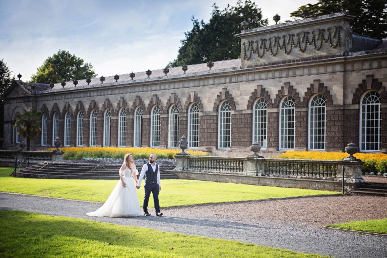 Elegant wedding photograph of bride and groom holding hands and walking down a path with the Orangery at Margam Country Park behind them.
