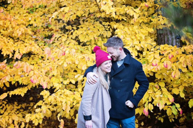 Colourful autumnal backdrop with couple hugging & smiling at Westonbirt Arboretum for their engagement shoot.