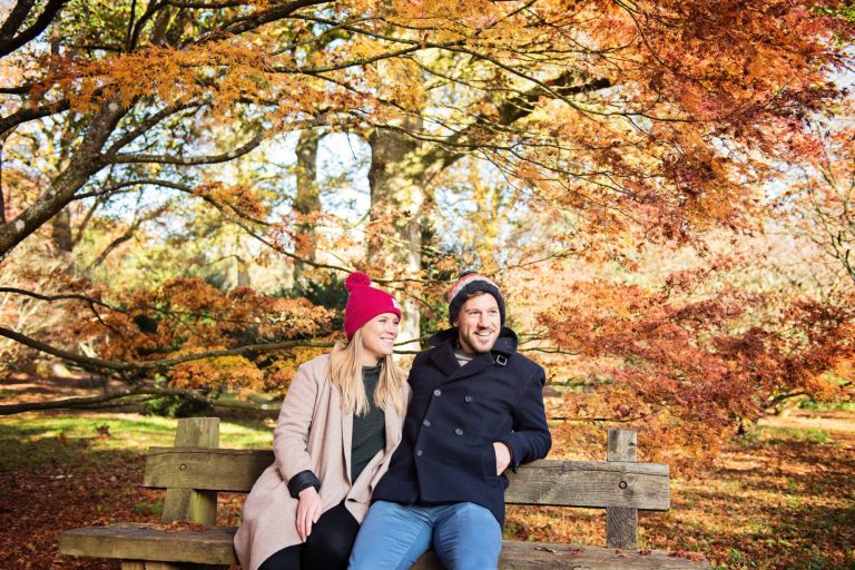 Colourful autumnal backdrop with couple smiling on a wooden bench at Westonbirt Arboretum for their engagement shoot.
