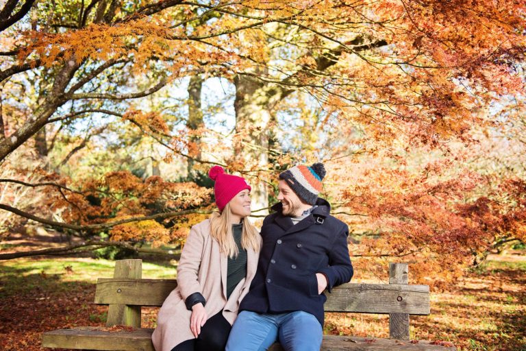 Colourful autumnal backdrop with couple smiling on a wooden bench at Westonbirt Arboretum for their engagement shoot.