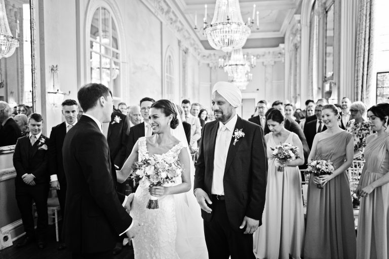 Photo of the bride meeting the groom for the first time after she has walked down the wedding isle at Danesfield House. Father of the bride and all guests look emotional. Black and white photograph.