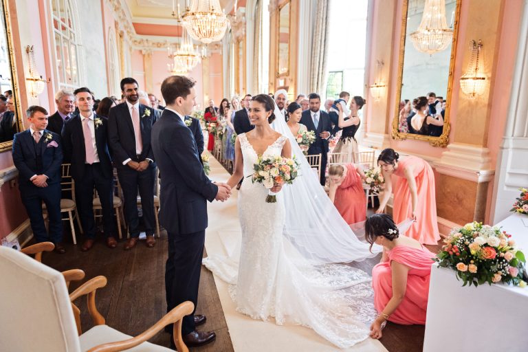 Bride and groom hold hands as they meet at the wedding aisle. Bridesmaids are spending time to correctly lay out the dress and veil.