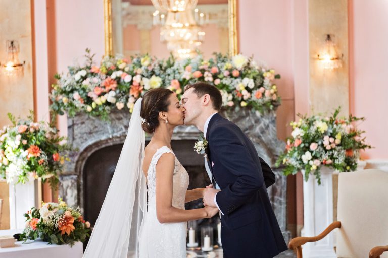 A bride and groom exchange a kiss after being announced "Husband and wife". A sea of stunning wedding flowers are behind them.