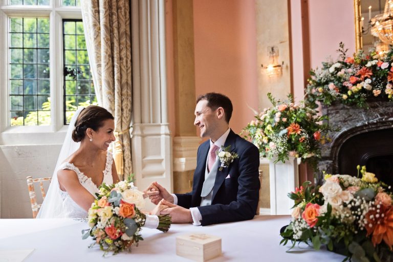 A bride and groom hold hands and smile and laugh at each other after signing the marriage certificate.