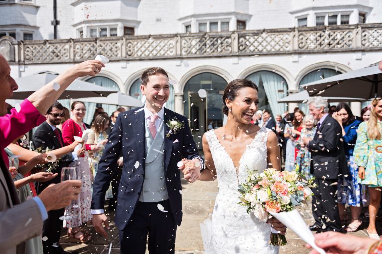 Photograph of the bride and groom (both smiling), walking down a confetti isle made by family and friends at Danesfield House Hotel and Spa.