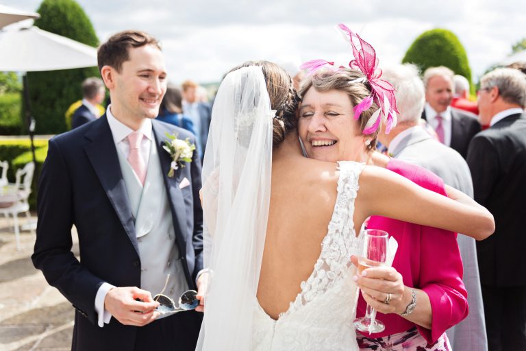 The mother of the groom hugs the bride with the groom onlooking. All smiling. Colourful candid photograph.