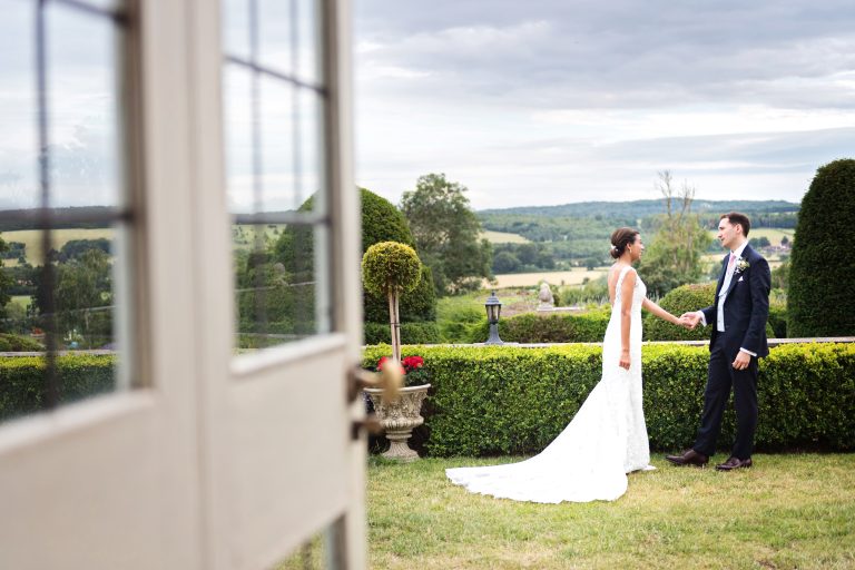 Bride and groom hold hands and have a moment together. Photographed candidly and composed as if looking through a window/door. Photographed at Danesfield House Hotel and Spa.