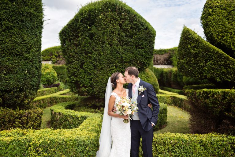 Bride and groom kiss with the magnificent topiary of Danesfield House behind them.