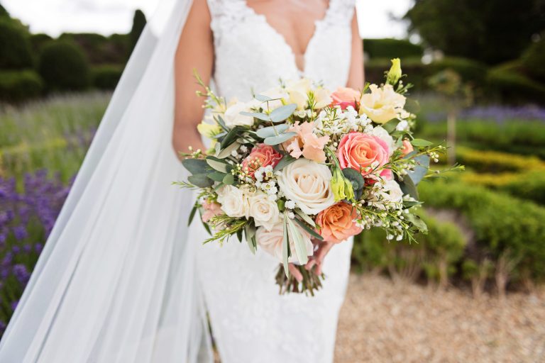 Mid centre photo of the bride, her wedding veil and wedding bouquet. The wedding bouquet is an elegant summer mix of roses, gypsophila and eucalyptus leaf. Photographed in the formal gardens of Danesfield House Hotel and Spa in the background.