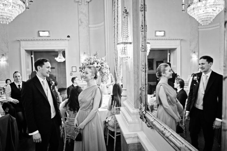 Groom chats with bride (times two) as image is doubled by using a mirror. Black and white. Chandelier in the ceiling. Danesfield House Ballroom.