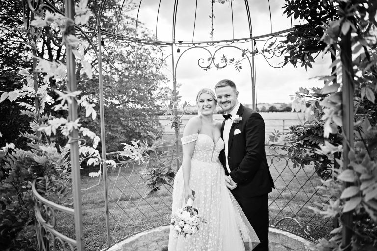 Candid photo B&W photo by Blooming Photography taken at Blackwell Grange, (Warwickshire) of a bride and groom smiling towards the camera whilst they are in a metal arch