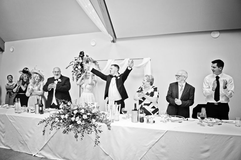 Black and white image of the groom with his hands in the air as the top table sign and clap with him. Candid photo by Blooming Photography. Taken at Blackwell Grange, Warwickshire.