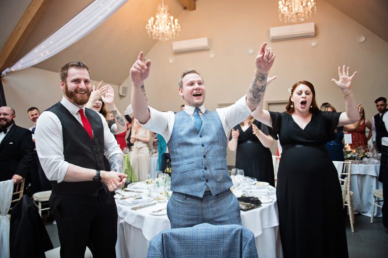 Image of wedding guests smiling and singing with their hands in the air during a song. Candid photo by Blooming Photography. Taken at Blackwell Grange, Warwickshire. Party!