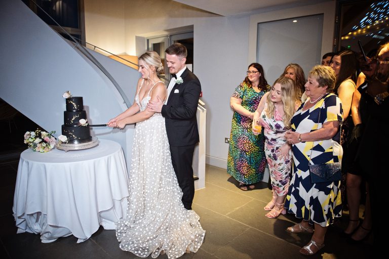 Candid photo by Blooming Photography. Taken at Blackwell Grange, Warwickshire. Image of bride and groom cutting the cake, guests look onwards.