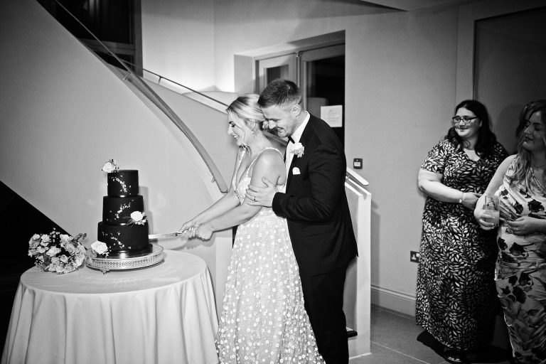 B&w image of bride and groom cutting the cake, guests look onwards. Candid photo by Blooming Photography. Taken at Blackwell Grange, Warwickshire.