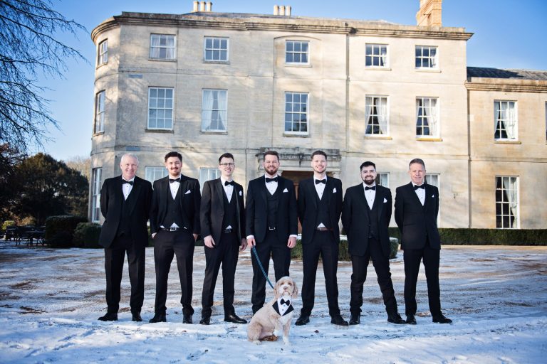 Seven groomsmen and a wonderful dog (dressed with a bowtie) stand tall in a line on snow with Eastington Park behind them.