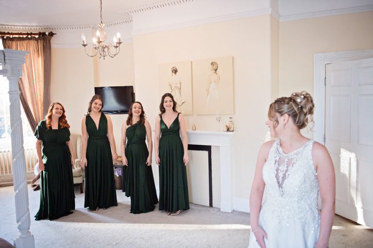 Bride looking over her shoulder at her bridesmaids, who are all smiling at the bride. Photo taken at Eastington Park by Blooming Photography