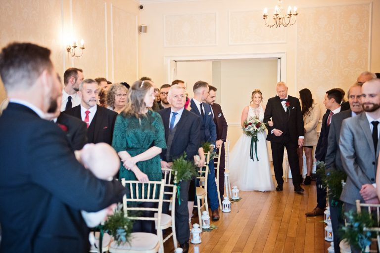 Bride walks down the wedding isle with her father in hand. Groom and rest of guests look onwards towards the bride.