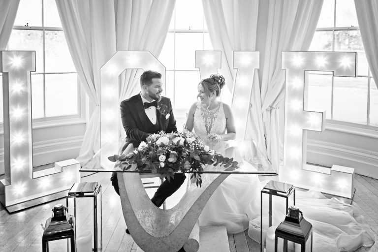 B&W image of Bride and groom as they are signing the wedding register in front of the bay windows at Eastington Park, with large lit up Love letters behind them.