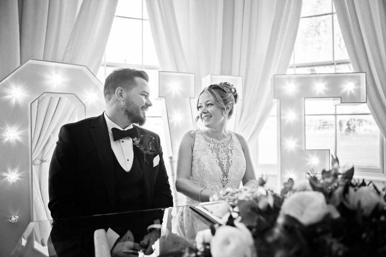 B&W image of Bride and groom as they are signing the wedding register in front of the bay windows at Eastington Park, with large lit up Love letters behind them.