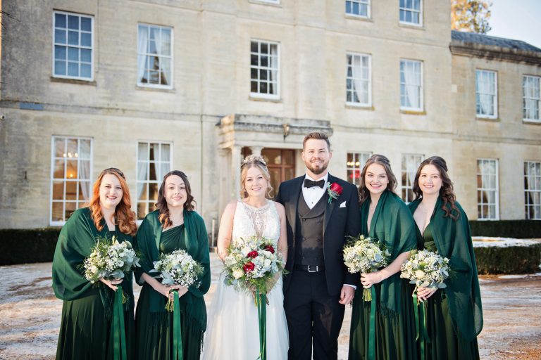 Bride and groom with bridesmaids standing in a line on snow with Eastington Park behind them. Looks beautiful.