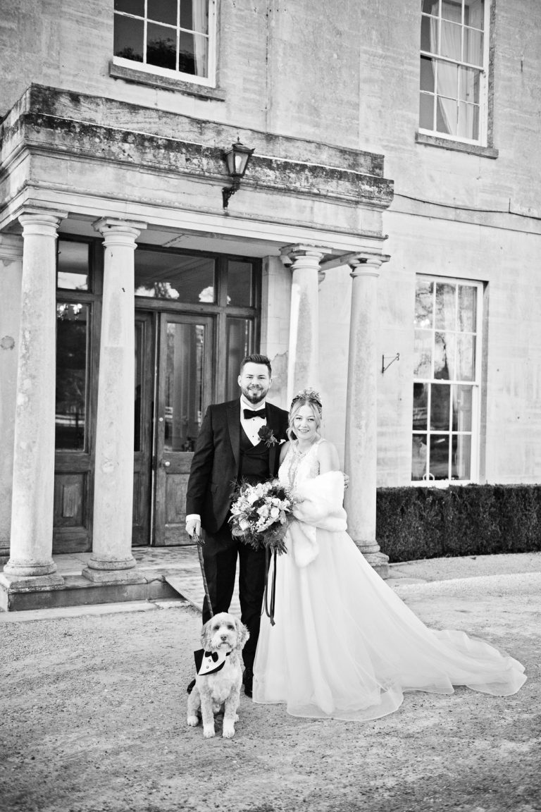 B&W photo of bride and groom standing with their dog outside the front of Eastington Park.