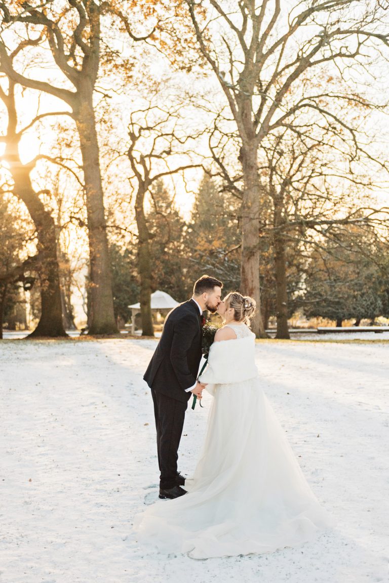 Winter snowy image of a bride and groom kissing together in the grounds of Eastington Park, with the sun setting behind them.