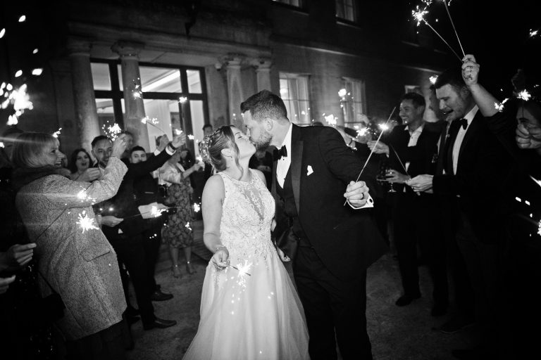 Bride and groom kiss with their friends and family surrounding them outside with sparklers. Black and white photo. Taken at Eastington Park at Christmas Wedding by Blooming Photography.