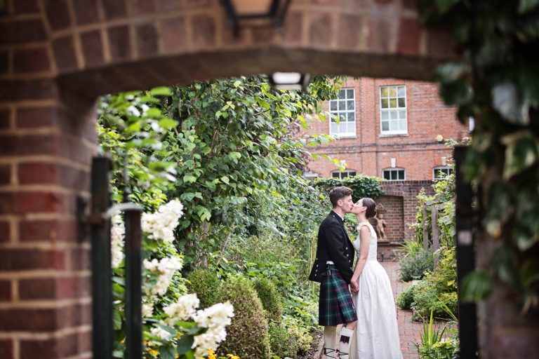 Candid wedding photo of bride and groom kissing through an arch at Hotel Du Vin Winchester, photo by Blooming Photography