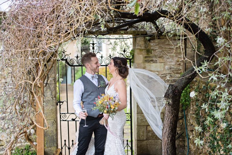 Bride and groom hang out under a climber and arch during a rain shower.