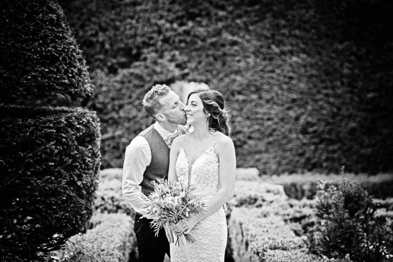 B&w photo of groom kissing bride on the cheek.