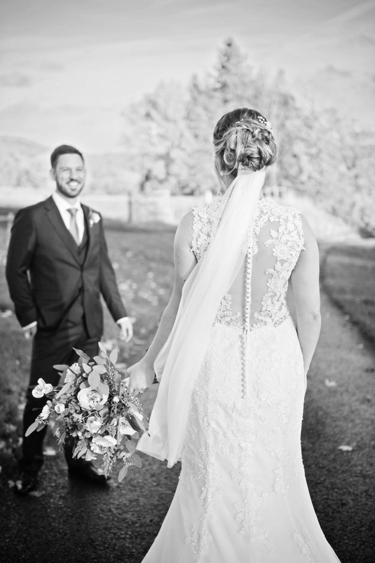 Black and white photograph of the back of a bride (with her beautiful wedding dress), with the groom smiling, facing her.