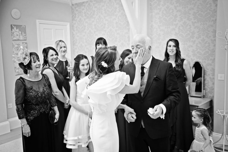 B&W storytelling image of the bride wiping away a tear from the father of the bride. The bridal party look on.